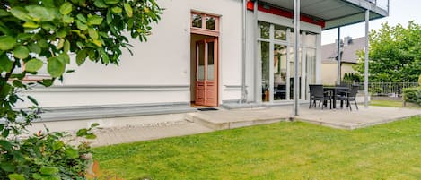 A residential building with a covered outdoor seating area, a red door, and a well-maintained lawn.