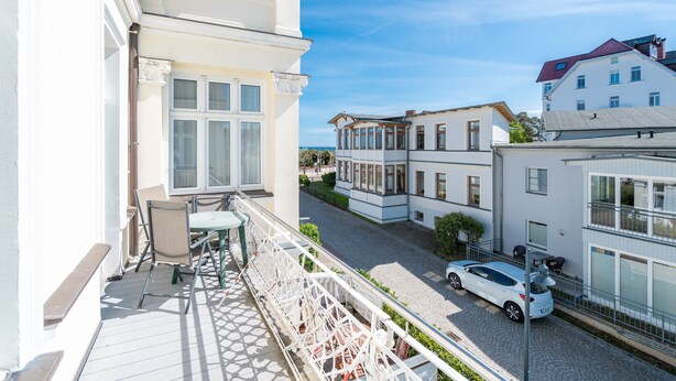A balcony with a table and chairs, a car parked on the street, and buildings in the background. - Villa Albertine, App. 07 (Heringsdorf)