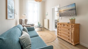 A living room with a sectional sofa, a wooden dresser, a television, and a large window with curtains. - Villa Albertine, App. 07 (Heringsdorf)