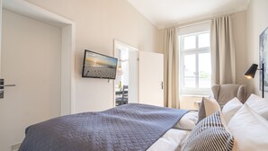 A neatly made bed with a blue quilt, a bedside lamp, and a window with curtains. - Villa Albertine, App. 07 (Heringsdorf)