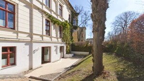 A two-story building with a white facade, red-framed windows, and a small garden area with a tree and shrubs. - Villa Arcadia, App. 10 (Heringsdorf)