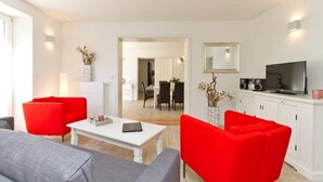 A modern living room with a white sofa, red chairs, a coffee table, and a TV on a white cabinet. - Villa Arcadia, App. 10 (Heringsdorf)
