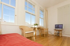 Ein Zimmer mit einem Bett, zwei Stühlen, einem kleinen Tisch mit einer Vase, einem Fernseher auf einem Schrank und großen Fenstern.