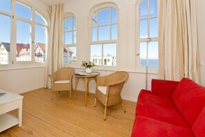 Une pièce dotée de grandes fenêtres en arc de cercle, d’un canapé rouge, de deux fauteuils en osier, d’une petite table ronde ornée d’un vase de fleurs et d’une table blanche.