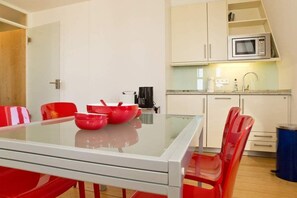 Een moderne keuken met witte kastjes, een magnetron en een spoelbak. Er staat een eettafel met rode stoelen en een glazen blad.