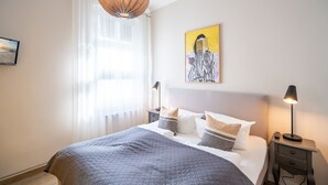 Ein ordentlich bezogenes Bett mit gesteppter Decke, zwei Nachttische mit Lampen, ein Gemälde an der Wand und ein Fenster mit transparenten Vorhängen.