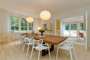 A spacious dining area with a wooden table and white chairs, large windows, and a view of the outdoors.