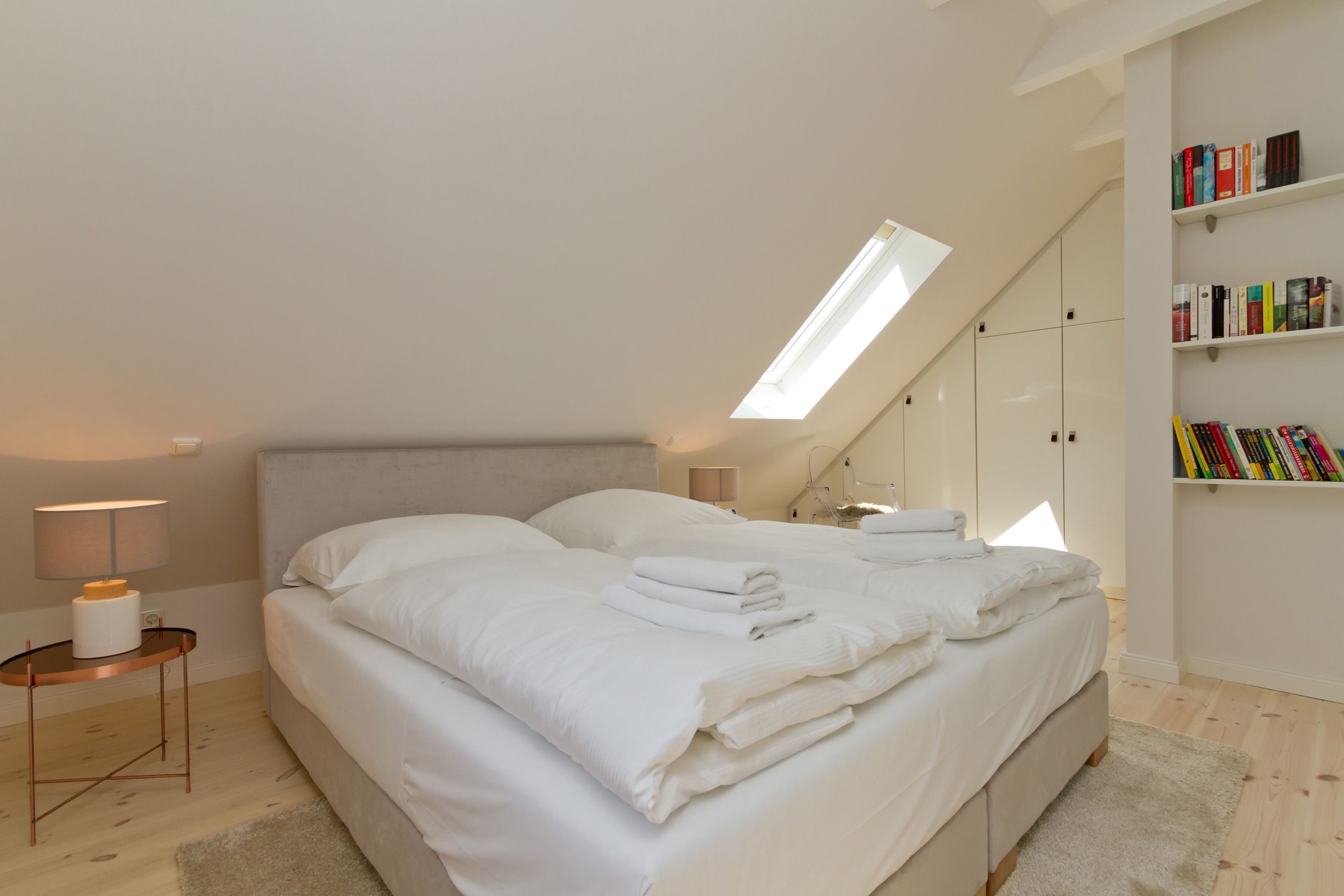 Ein Schlafzimmer mit einem großen Bett, einem Nachttisch mit Lampe, einem Oberlicht und einem Bücherregal mit Büchern.
