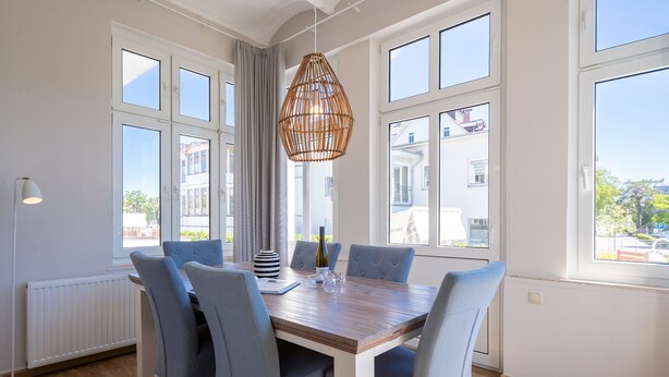 Comedor con mesa de madera, sillas tapizadas en gris, lámpara colgante de mimbre y ventanas amplias que ofrecen vistas a edificios y vegetación.