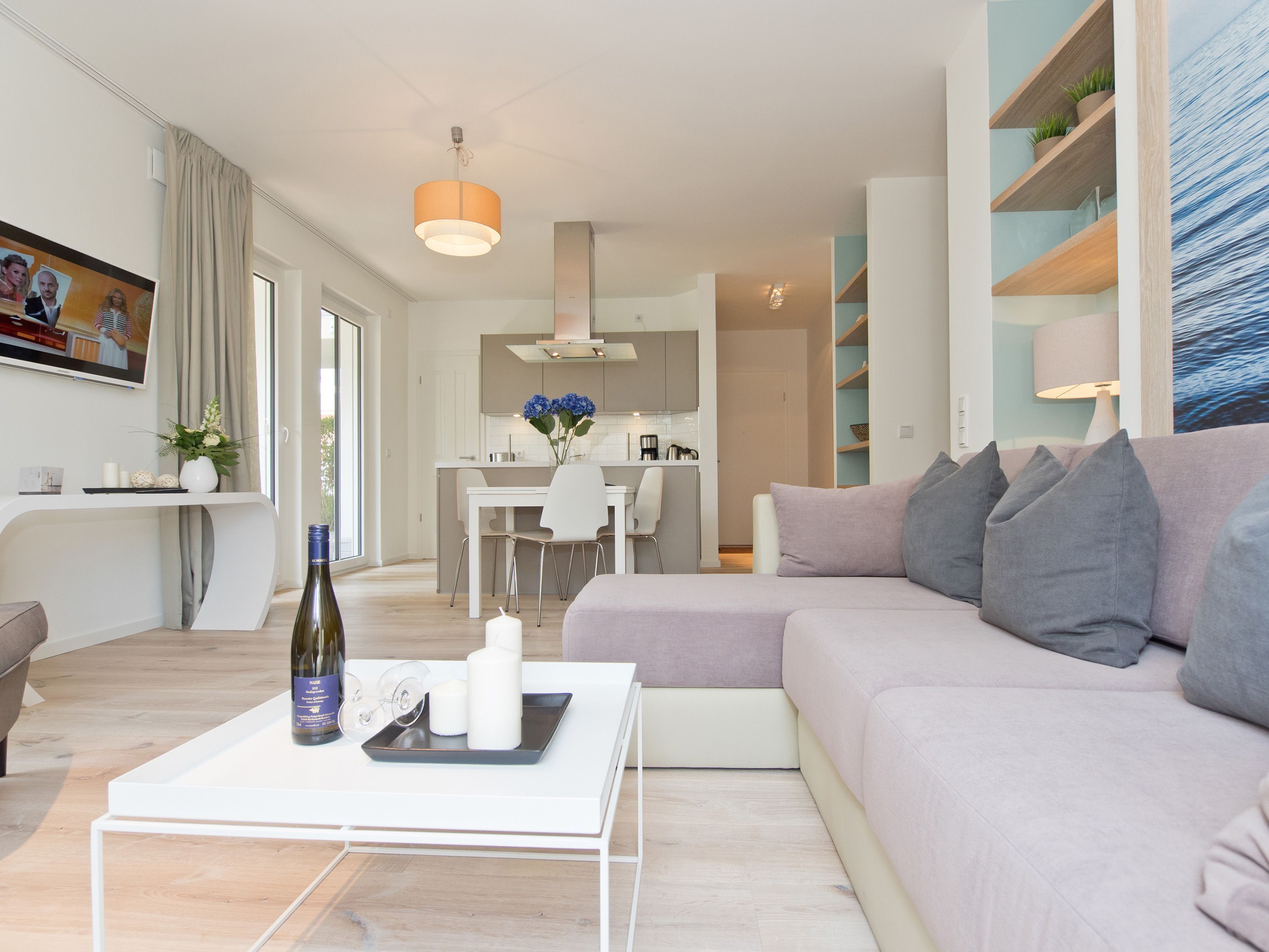 A modern living room with a sofa, a coffee table with a bottle and glasses, a dining area with a table and chairs, and a kitchen in the background.