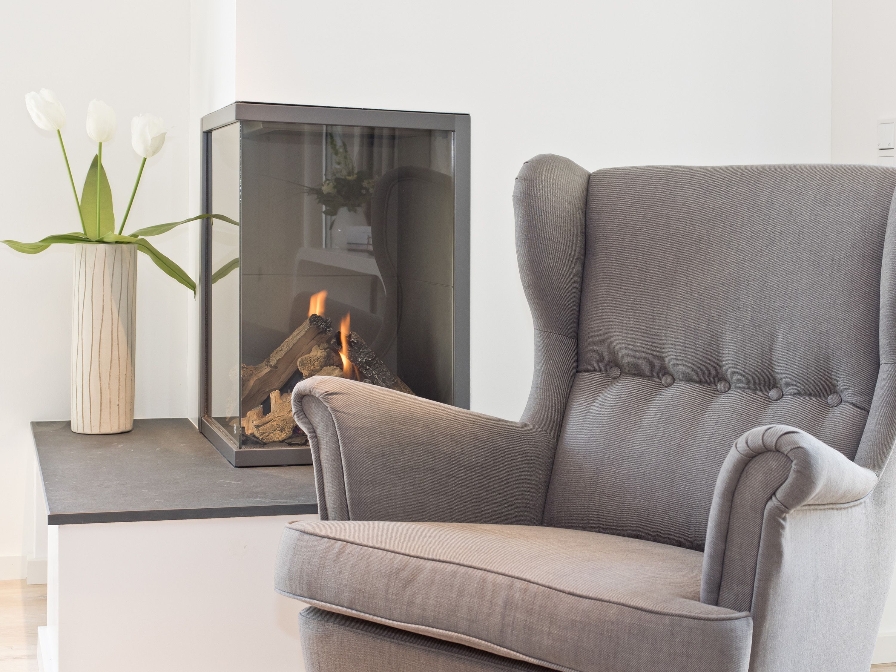 A modern living room with a grey upholstered armchair, a glass fireplace with a visible fire, and a white vase with white flowers on a side table.
