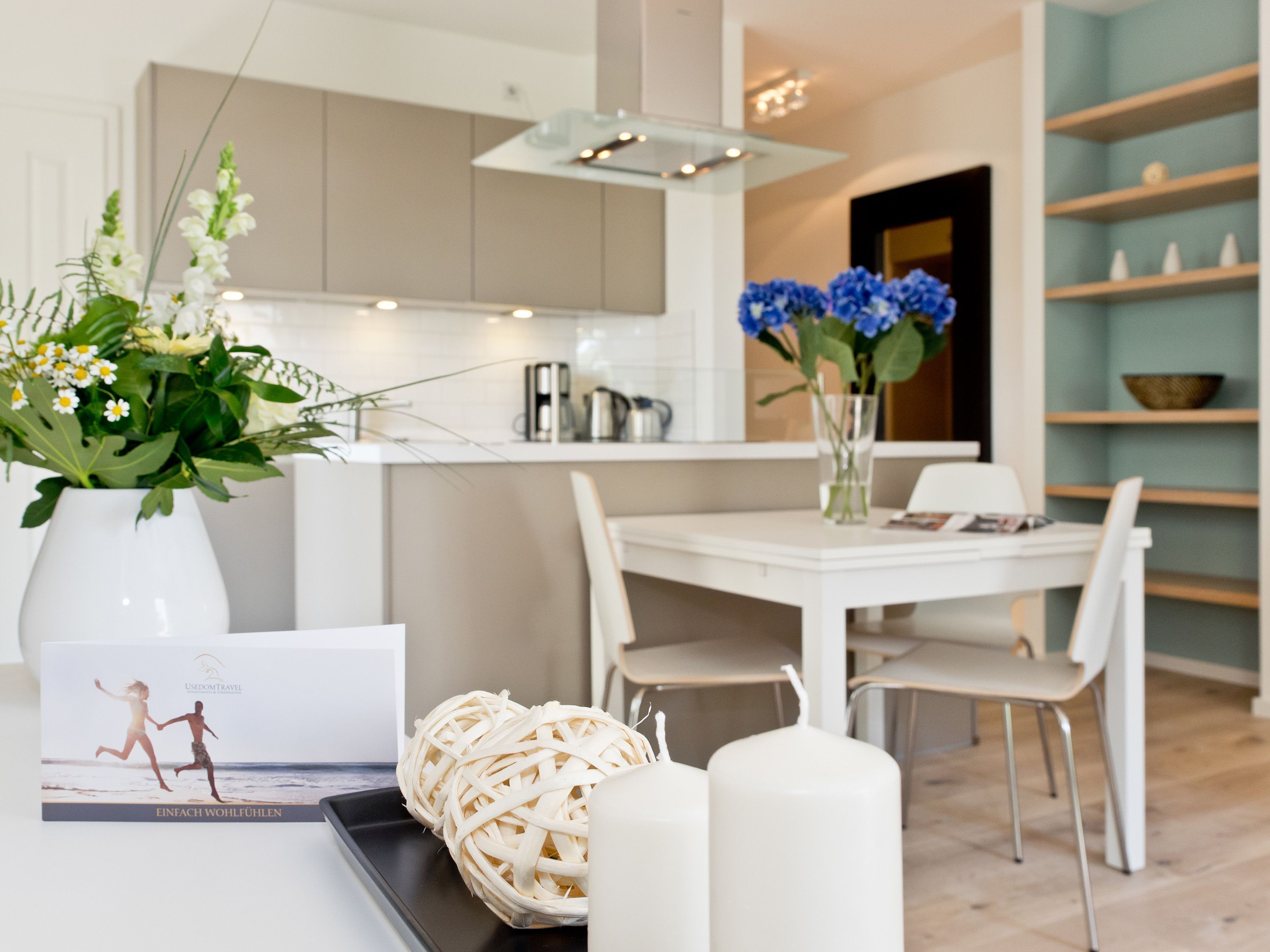 Una cucina moderna con mobili bianchi, un'area pranzo con tavolo e sedie bianche e un vaso con fiori blu sul tavolo.
