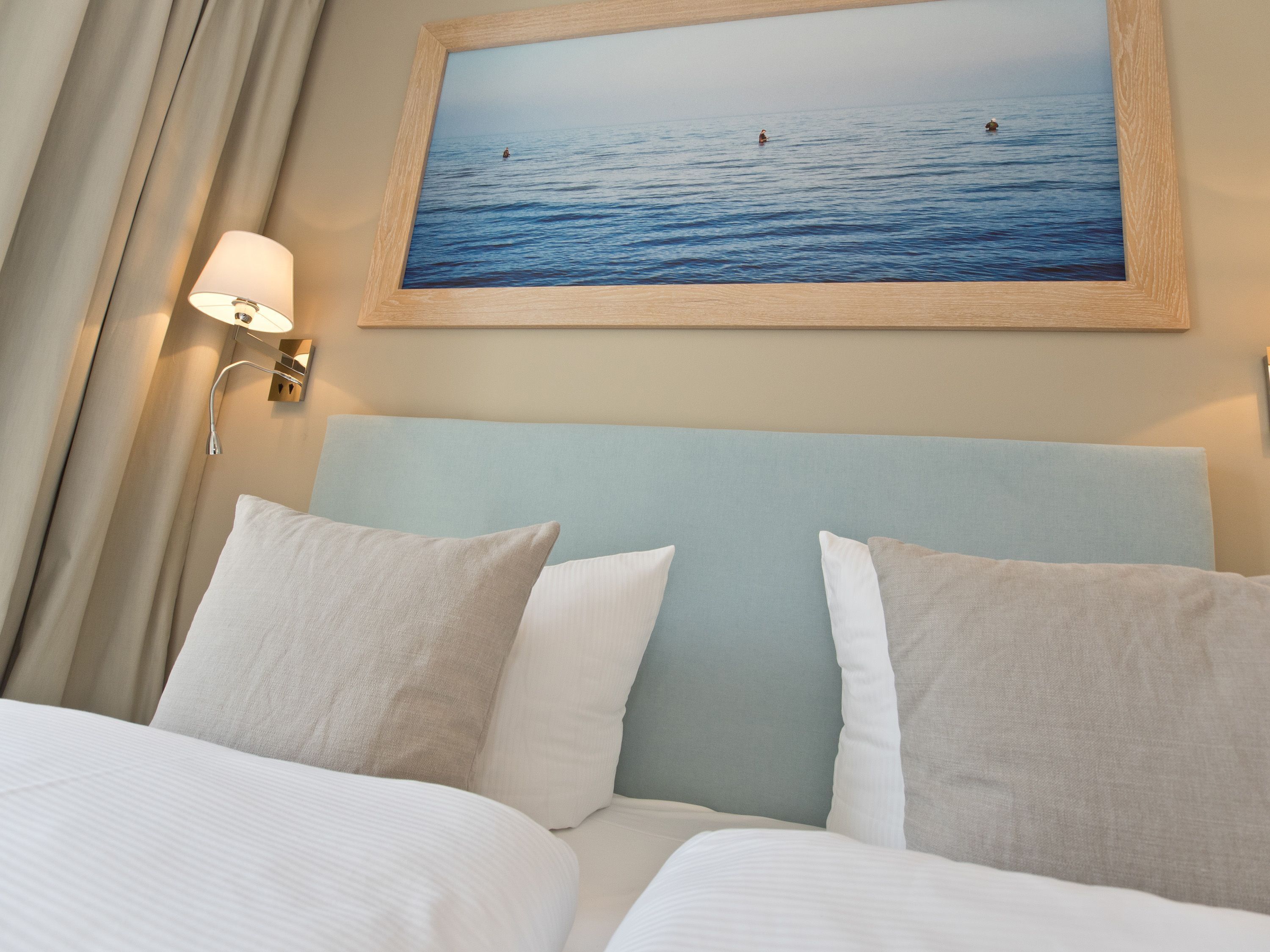 Hotelkamer met een bed, kussens, een hoofdeinde, een wandlamp en een lijst met een afbeelding van de zee.
