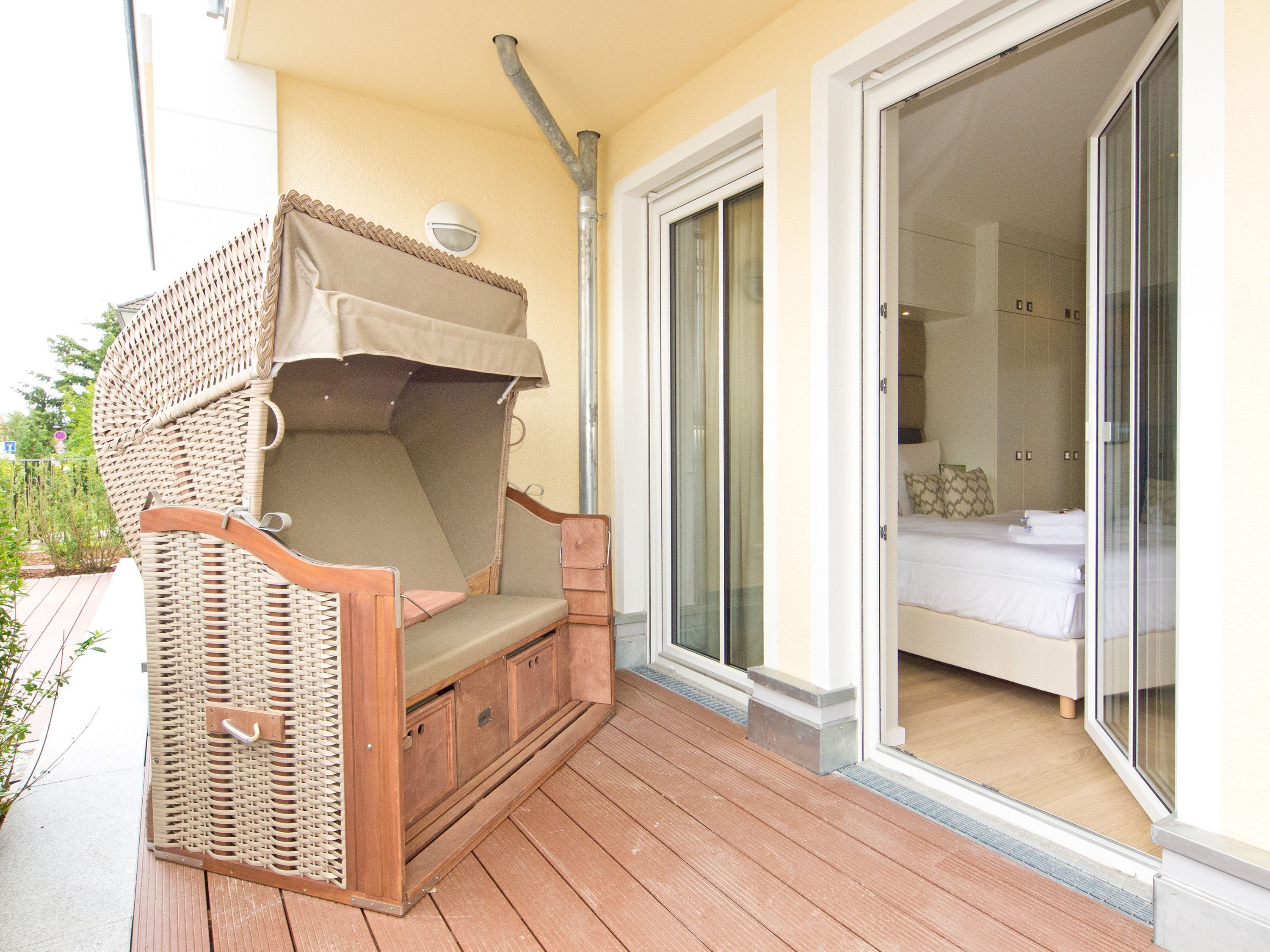 Un espace extérieur aménagé avec des sièges en bois, comprenant une chaise rembourrée et un auvent, situé à côté d’une porte-fenêtre coulissante qui donne sur une chambre avec un lit et une table de chevet.