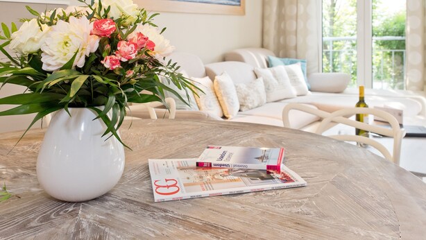 En stue med en hvid vase med blomster på et træbord, et magasin og en sofa med puder.