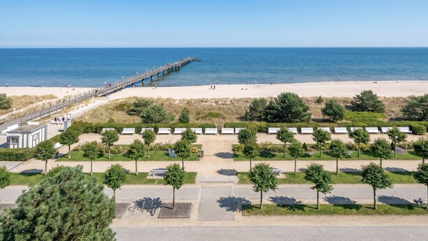 En strand med en bro, en række strandhytter og et parkeringsområde med træer.