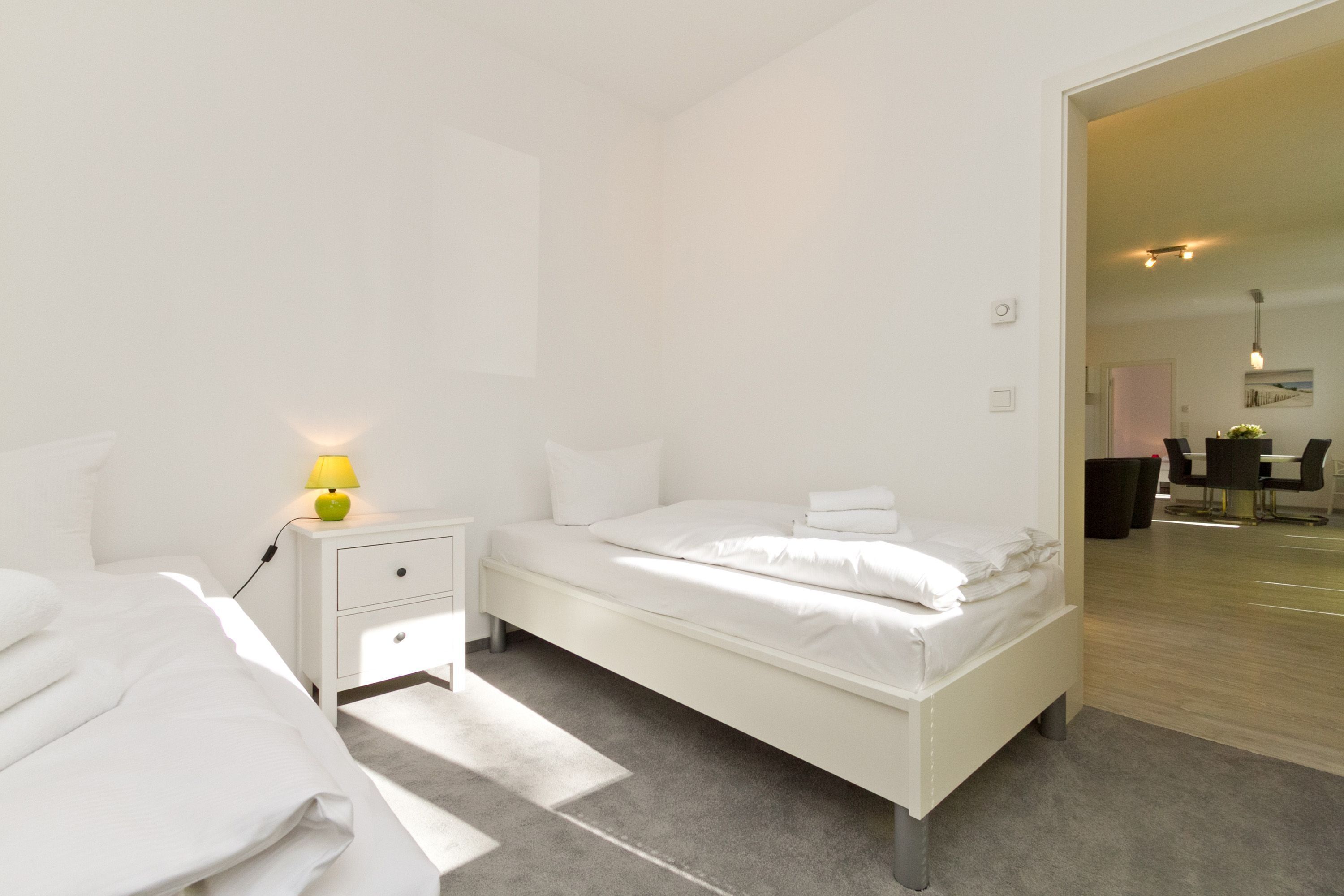 Ein modernes Schlafzimmer mit einem weißen Bett, einem Nachttisch mit Lampe und Blick in einen Essbereich.
