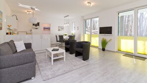 A modern living room with a grey sofa, white coffee table, and black chairs. There is a kitchen area with white cabinets and a TV. Large windows offer a view of trees. - Residenzen am Kulm, Villa Dorian 10 (Heringsdorf)