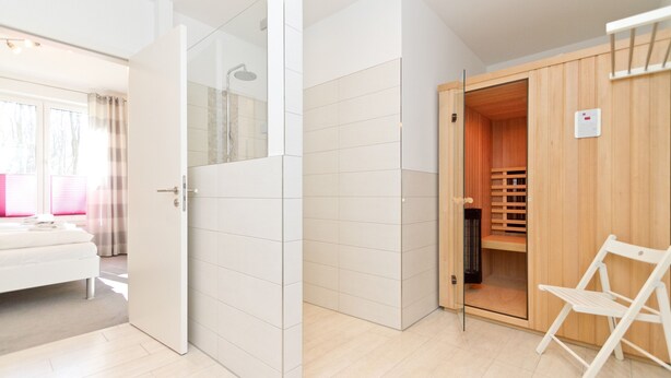 A modern hotel room with a sliding glass door leading to a shower area, a wooden closet, and a white chair. - Residenzen am Kulm, Villa Dorian 10 (Heringsdorf)