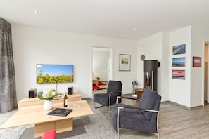 Ein modernes Wohnzimmer mit Holztisch, Flachbildfernseher, einer Couch und einem Esstisch mit Stühlen.