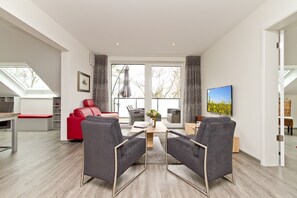 Ein modernes Wohnzimmer mit einem roten Sofa, grauen Sesseln, einem Holztisch und einem Fernseher an der Wand.