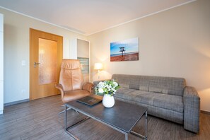 A modern living room with a leather armchair, a grey sofa, a coffee table with a laptop and a vase of flowers, a wooden door, and a painting of a pier on the wall.