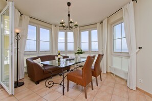 Een eetruimte met een glazen tafel, een bruine leren bank en vier stoelen. Er zijn grote ramen met witte gordijnen, een kroonluchter en uitzicht op zee.