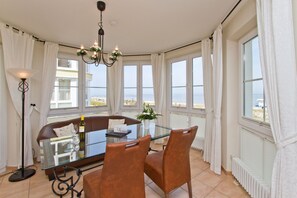 Un coin repas avec une table en verre, des chaises marron, un lustre et de grandes fenêtres avec des rideaux.