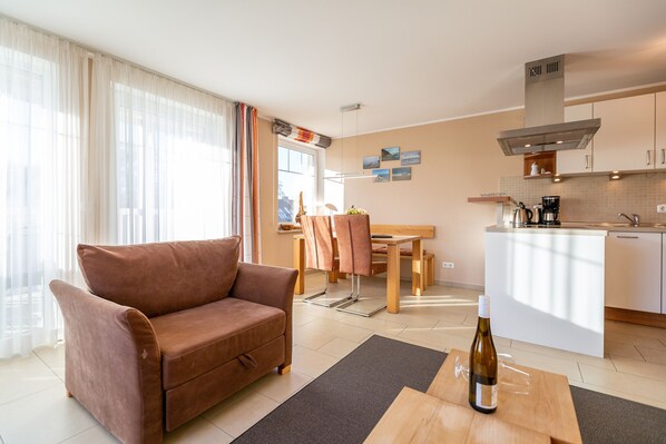 A modern living room with a brown sofa, a dining area with a wooden table and chairs, and a kitchen with a white island and stainless steel appliances.