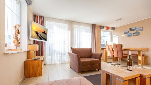 A modern hotel room with a bed, a wooden desk, a chair, a TV displaying a sunset scene, and a wooden side table with a decorative item.