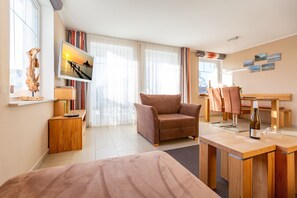 A modern hotel room with a bed, a wooden desk, a chair, a TV displaying a sunset scene, and a wooden side table with a decorative item.