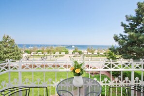 Un balcon avec une balustrade blanche, une table ornée d’un vase de fleurs, et une vue sur la mer et un parc.