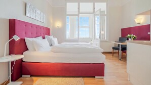 Habitació d'hotel moderna amb un llit tapissat de color vermell, roba de llit blanca, un escriptori amb una cadira i una finestra gran que aporta llum natural.