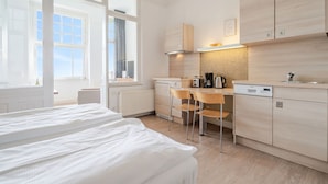 A modern hotel room with a bed, a small kitchenette, and a view of the sky.