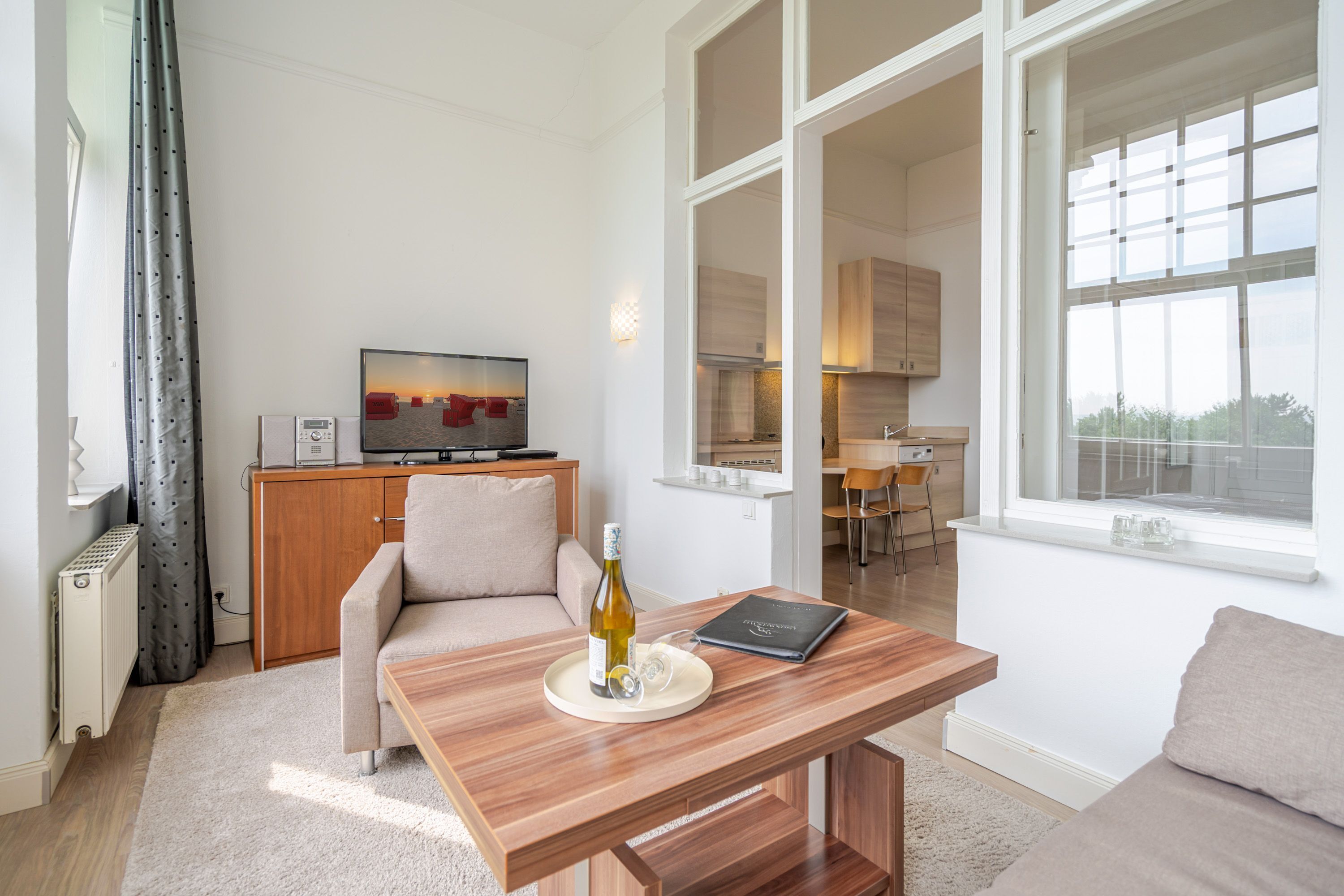 A modern living room with a sofa, a wooden coffee table with a bottle and glasses, a TV, and a view of the kitchen and outside.