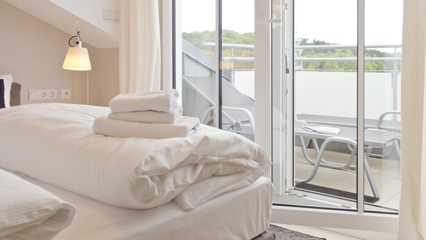 Habitación de hotel con cama, lámpara de pie y vista a un balcón con mobiliario exterior.