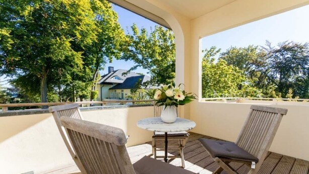 En balkon med to træstole, et lille rundt bord, en vase med blomster og udsigt til træer og et hus.