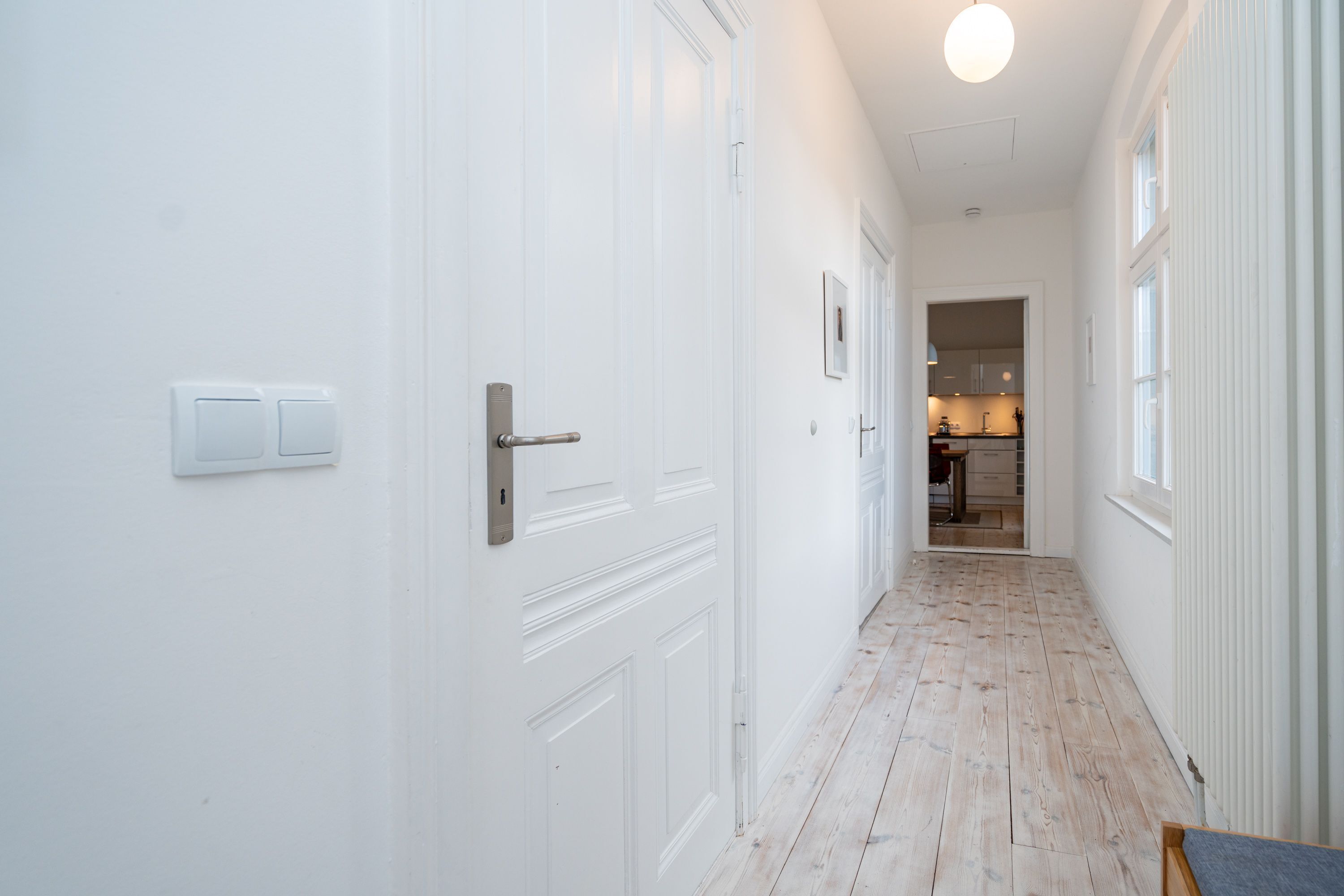 A hallway with white doors, a wooden floor, and a kitchen visible in the distance.