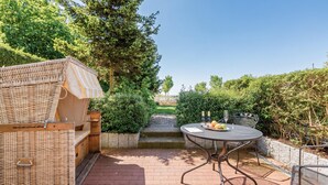 Une terrasse avec un canapé en osier, une table ronde en métal entourée de chaises, et un petit escalier menant à une zone herbeuse.