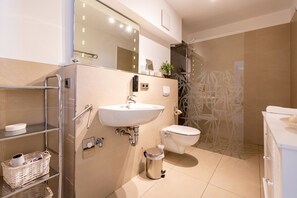 A modern bathroom with a white sink, toilet, and shower. There are shelves with baskets and a small trash can.