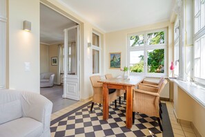 A dining area with a checkered floor, a wooden table, wicker chairs, and a view of greenery through large windows. - The Villa, App. 9, Germany (Heringsdorf)