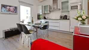 Una cocina moderna con armarios blancos, electrodomésticos de acero inoxidable y una zona de comedor con mesa de vidrio y sillas negras.