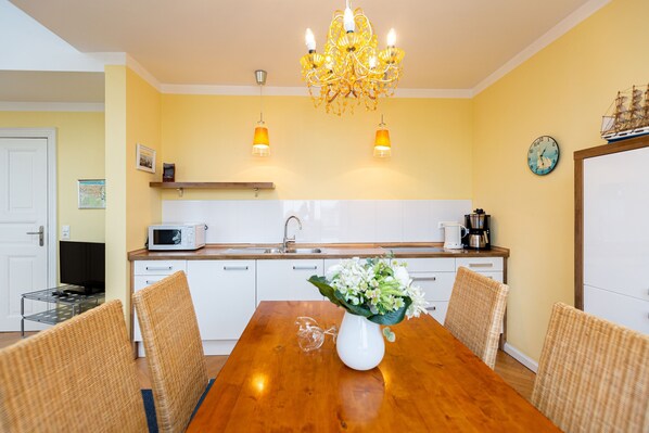 Un coin repas avec une table en bois, des chaises en osier, un vase blanc rempli de fleurs, un lustre, un four à micro-ondes et une machine à café posés sur le comptoir.