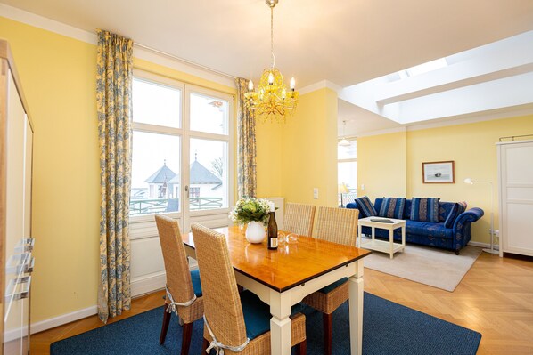 Ein Essbereich mit Holztisch, Stühlen und einer Chandelier. Im Hintergrund befindet sich eine Couch und ein Fenster mit Vorhängen.