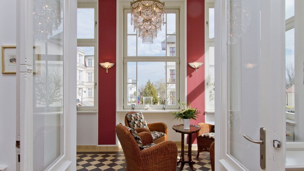 A room with a checkered floor, a wicker chair, a small table with a plant, and a window with a view of buildings.