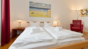 A hotel room with two beds, a red chair, a painting of a beach scene, and a lamp on each side of the beds.