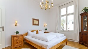 Ein Schlafzimmer mit einem hölzernen Bett, weißer Bettwäsche, zwei Nachttischen mit Lampen, einer Chandelüster, einem Fenster mit Vorhängen und einem Holzschrank.