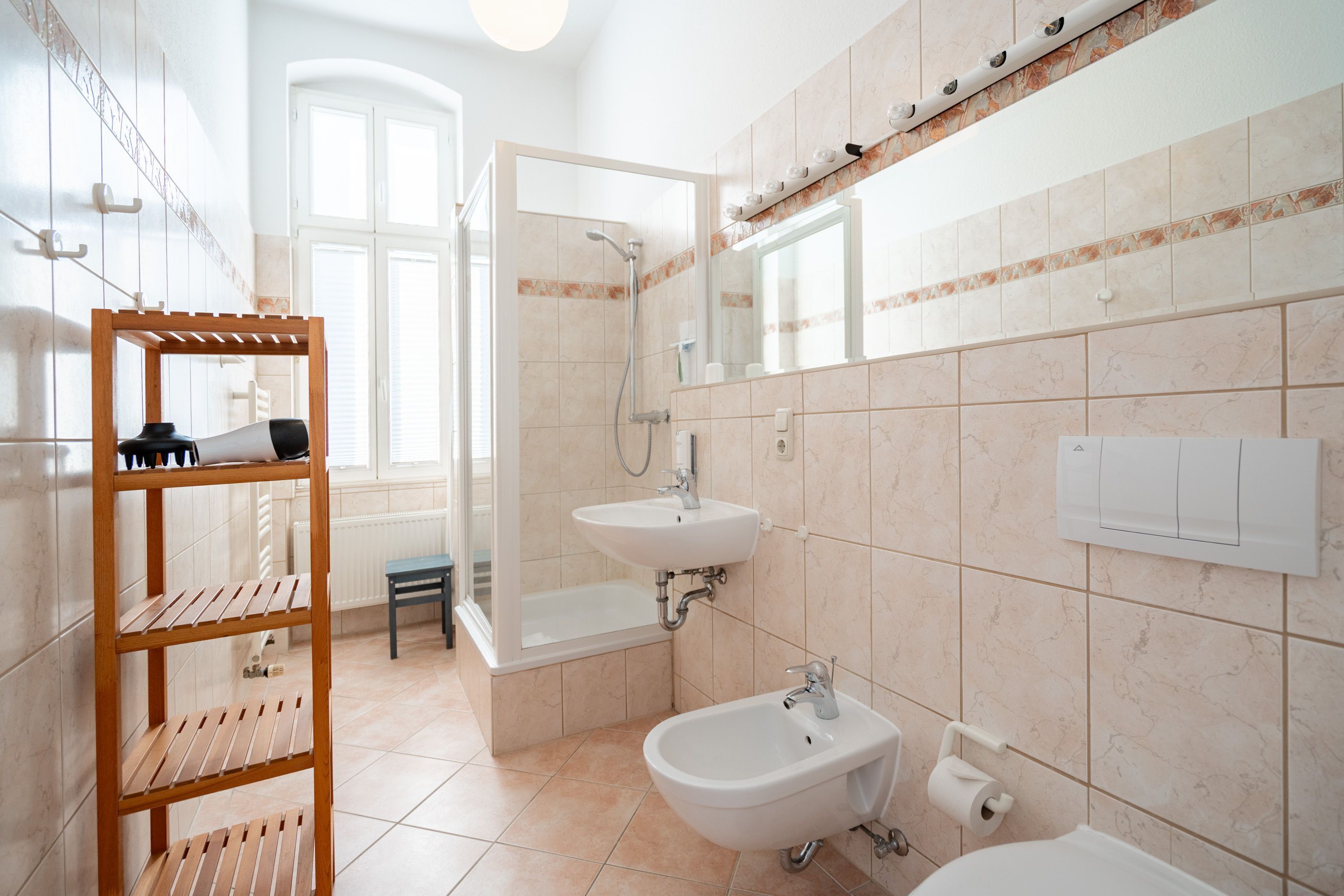 A modern bathroom with a shower, sink, and bidet. There is a wooden shelf with toiletries and a small table.