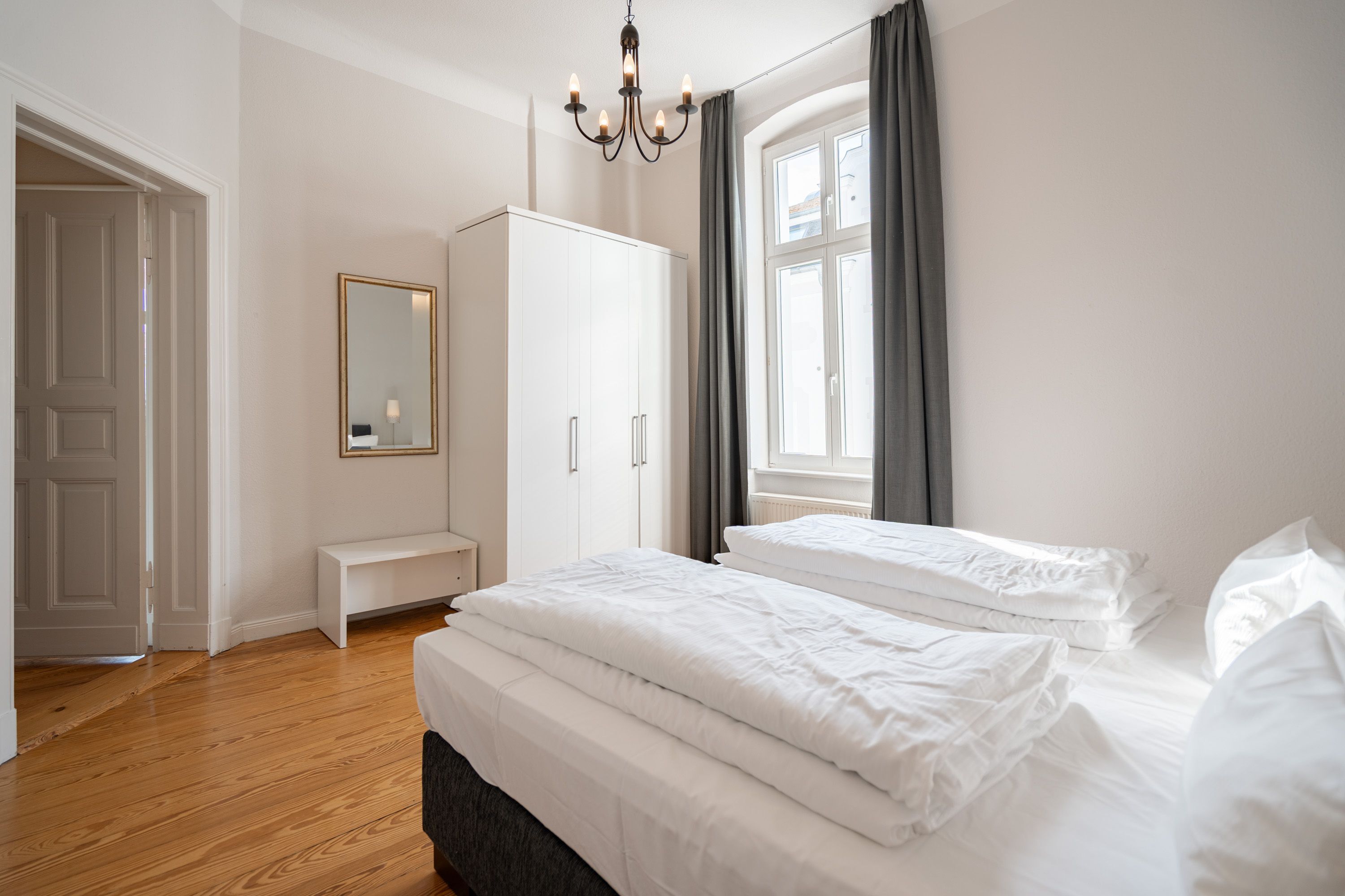 A bedroom with two beds, a wardrobe, a bench, and a large window with curtains.