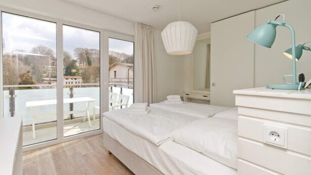 A bedroom with a large bed, a desk, a chair, and a balcony with a view of the outside.